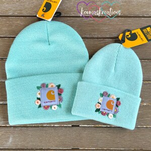 May include: Two turquoise beanies with embroidered floral designs and the Carhartt logo. One beanie is larger than the other. Both beanies have a folded brim. The background is a wooden surface.