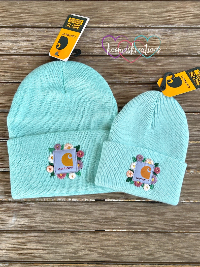 May include: Two turquoise beanies with embroidered floral designs and the Carhartt logo. One beanie is larger than the other. Both beanies have a folded brim. The background is a wooden surface.