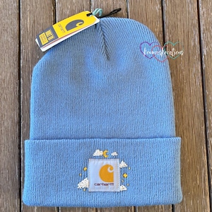 May include: A light blue beanie with a yellow Carhartt logo and embroidered clouds and stars. The beanie has a fold-over cuff.