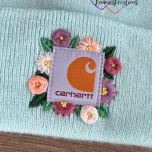 May include: A light blue knit beanie featuring a Carhartt logo patch surrounded by embroidered flowers in shades of pink, purple, and white. The beanie has a ribbed texture and a folded brim. The logo is orange and the text "Carhartt" is in red.