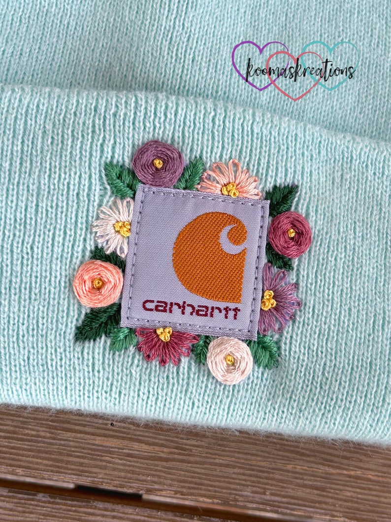 May include: A light blue knit beanie featuring a Carhartt logo patch surrounded by embroidered flowers in shades of pink, purple, and white. The beanie has a ribbed texture and a folded brim. The logo is orange and the text "Carhartt" is in red.