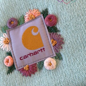 May include: A light blue knit beanie featuring an embroidered floral design around a Carhartt logo patch. The flowers are in shades of pink, purple, and peach, with green leaves. The Carhartt logo is mustard yellow on a gray patch, with the brand name in burgundy.