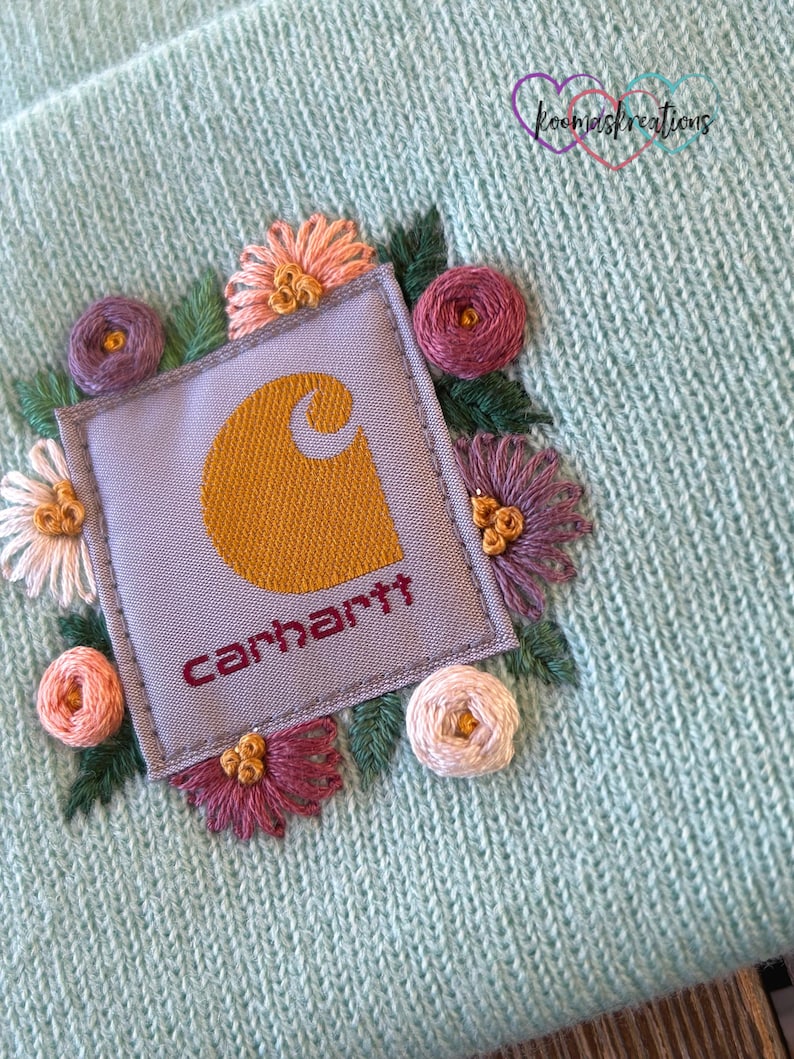 May include: A light blue knit beanie featuring an embroidered floral design around a Carhartt logo patch. The flowers are in shades of pink, purple, and peach, with green leaves. The Carhartt logo is mustard yellow on a gray patch, with the brand name in burgundy.