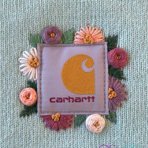 May include: A light blue knit beanie featuring an embroidered Carhartt logo patch surrounded by colorful floral embroidery. The Carhartt logo is in gold with the brand name in burgundy. The flowers are in shades of pink, purple, and white.
