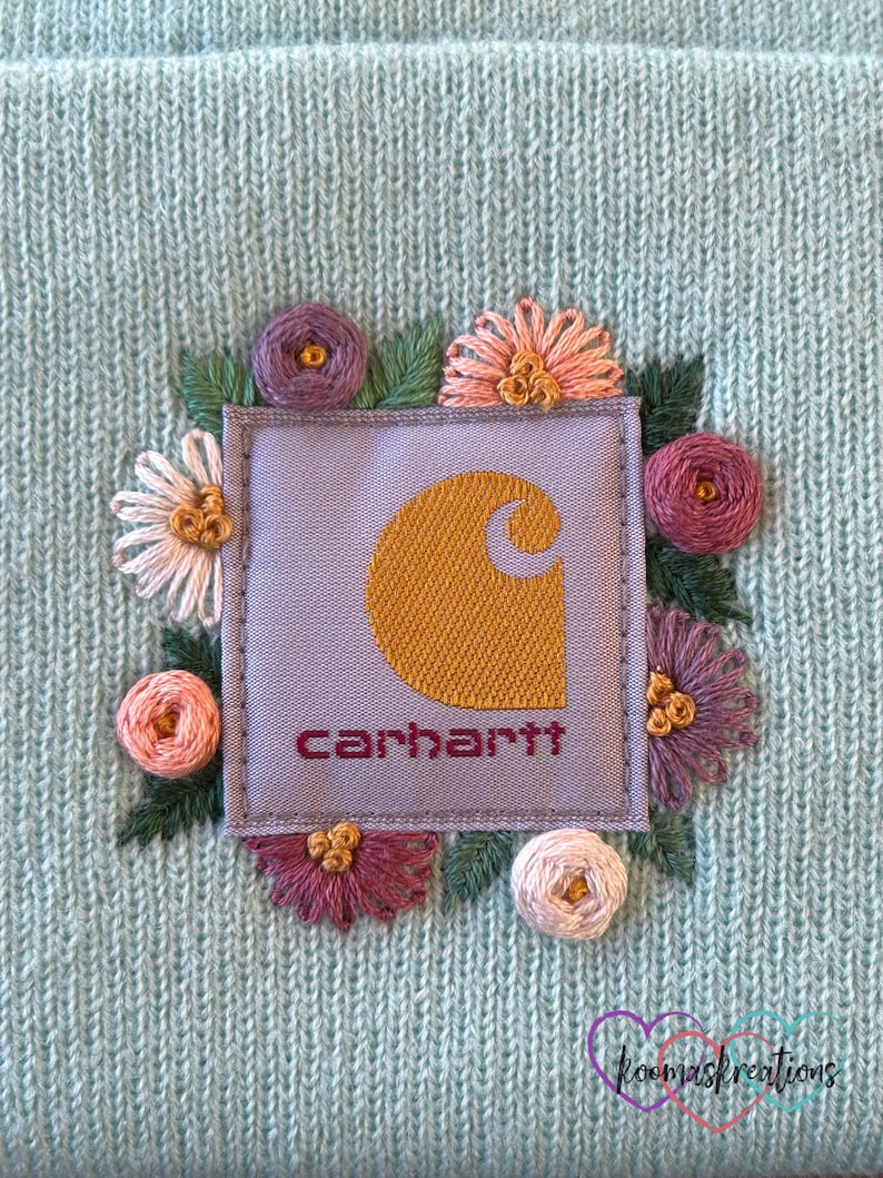 May include: A light blue knit beanie featuring an embroidered Carhartt logo patch surrounded by colorful floral embroidery. The Carhartt logo is in gold with the brand name in burgundy. The flowers are in shades of pink, purple, and white.