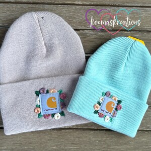 May include: Two knit beanies, one light gray and one aqua blue, are displayed. Each beanie features a square patch with the Carhartt logo and floral embroidery. The beanies are folded at the brim and rest on a wooden surface.