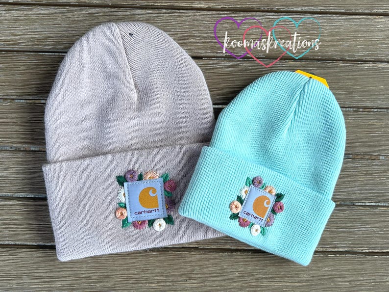 May include: Two knit beanies, one light gray and one aqua blue, are displayed. Each beanie features a square patch with the Carhartt logo and floral embroidery. The beanies are folded at the brim and rest on a wooden surface.