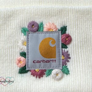 May include: Cream-colored beanie featuring an embroidered patch with the Carhartt logo in gold and the brand name in burgundy. The patch is surrounded by colorful embroidered flowers in pink, purple, and white, with green leaves. The beanie has a knitted texture.