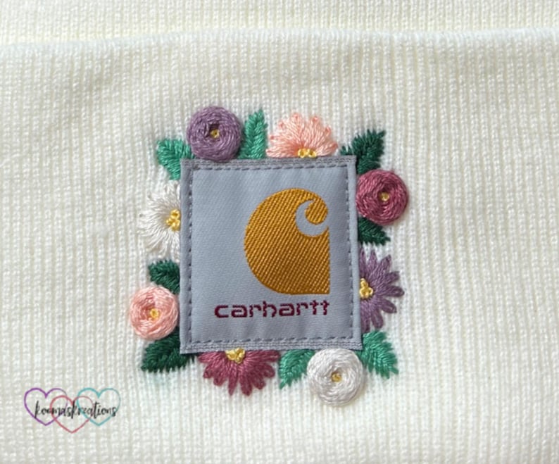May include: Cream-colored beanie featuring an embroidered patch with the Carhartt logo in gold and the brand name in burgundy. The patch is surrounded by colorful embroidered flowers in pink, purple, and white, with green leaves. The beanie has a knitted texture.