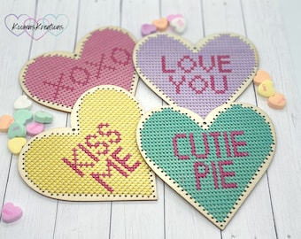 Peut inclure: Quatre ornements en bois en forme de cœur avec des motifs de point de croix. Les ornements sont décorés des phrases "XOXO", "LOVE YOU", "SO KISS ME" et "CUTIE PIE".