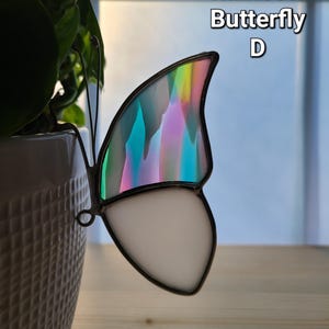 May include: A stained glass butterfly with a rainbow wing and a white lower wing section. The butterfly is attached to a white ceramic pot with a plant. The text "Rainbow Butterfly D" is in the upper right corner.