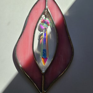 Puede incluir: Un atrapasol de vidrio tintado de color rosa con un cristal arco&iacute;ris colgando en el centro. El atrapasol tiene forma de coraz&oacute;n con la parte inferior puntiaguda.