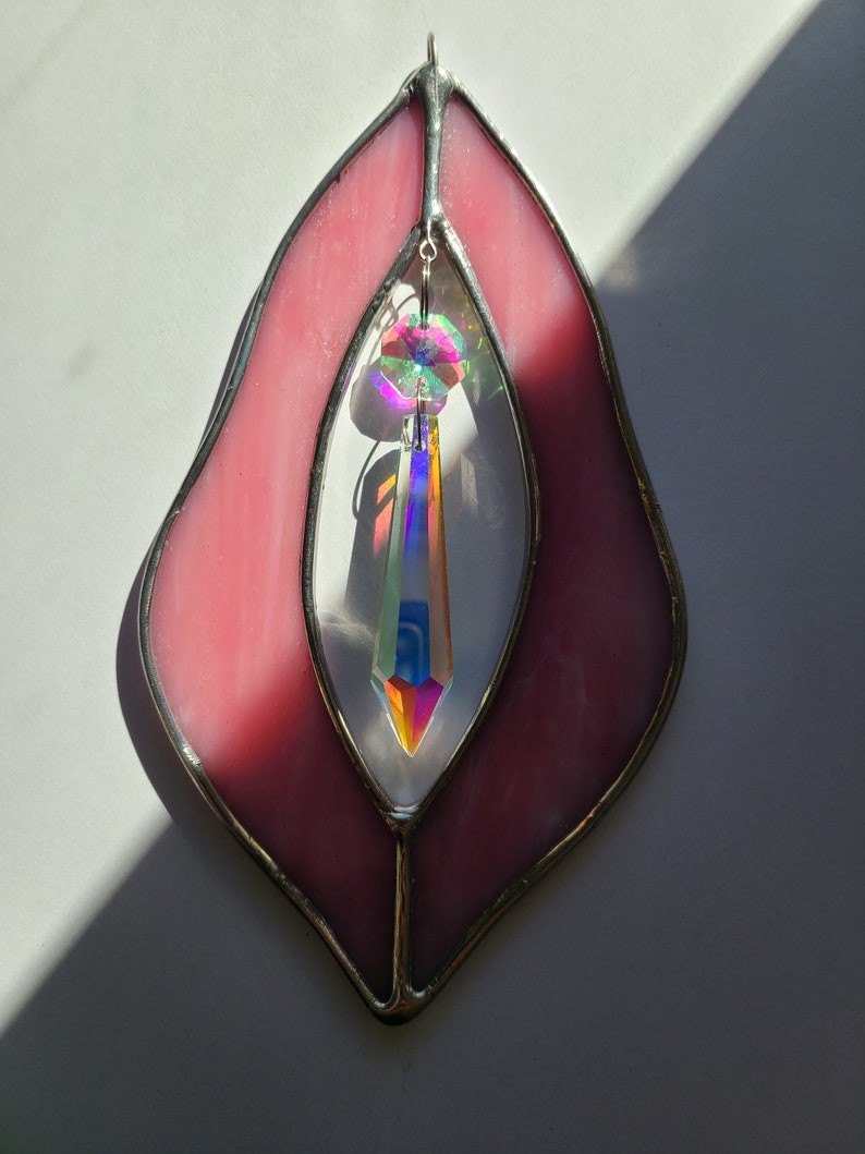 Puede incluir: Un atrapasol de vidrio tintado de color rosa con un cristal arco&iacute;ris colgando en el centro. El atrapasol tiene forma de coraz&oacute;n con la parte inferior puntiaguda.