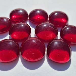 Puede incluir: Una colección de diez piedras de vidrio rojas, lisas y redondeadas. Las piedras tienen un acabado brillante y un color profundo y rico, con un tono ligeramente más claro en la parte superior. Están dispuestas sobre una superficie blanca.