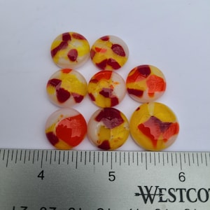 Puede incluir: Nueve cabujones redondos de vidrio en amarillo, rojo y blanco. Los cabujones están sobre una superficie blanca junto a una regla. Cada cabujón tiene aproximadamente 2,5 cm de diámetro.