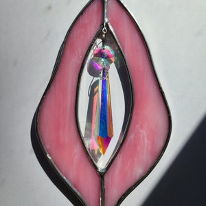 Puede incluir: Ventana de cristal de color rosa con un prisma de cristal colgando en el centro. La ventana tiene forma de l&aacute;grima y un marco plateado.