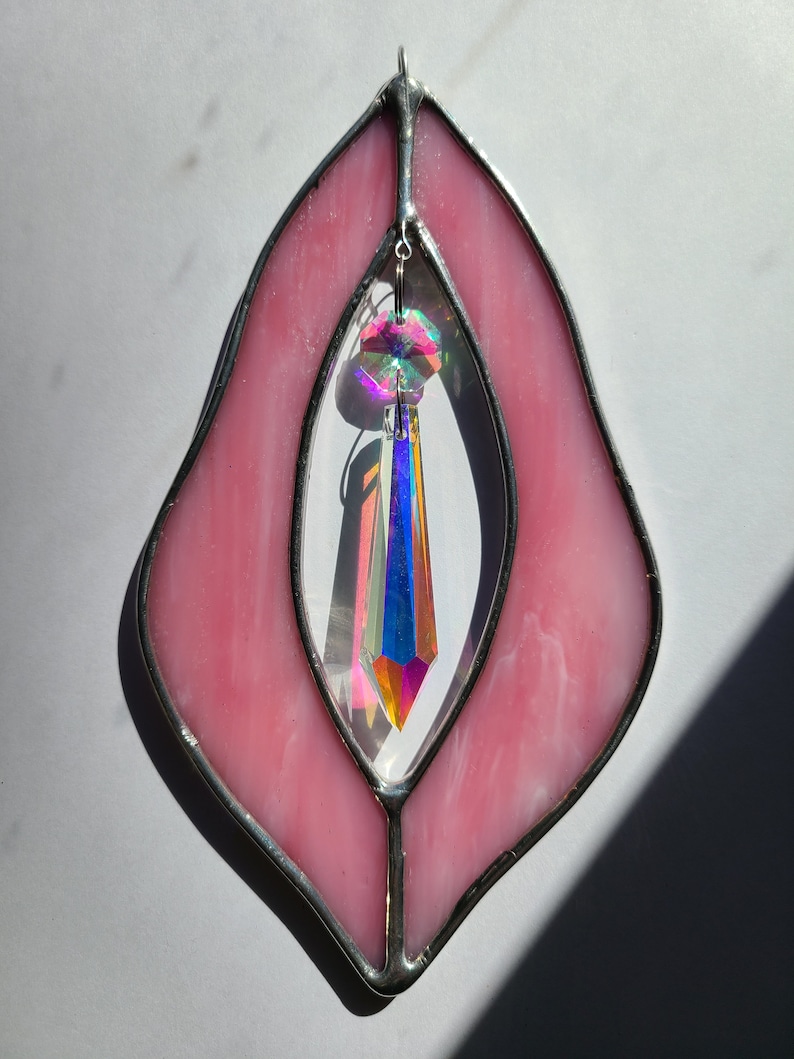 Puede incluir: Ventana de cristal de color rosa con un prisma de cristal colgando en el centro. La ventana tiene forma de l&aacute;grima y un marco plateado.
