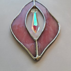 Puede incluir: Una pieza de vidrio coloreado rosa en forma de hoja con un cristal transparente iridiscente colgando en el centro.