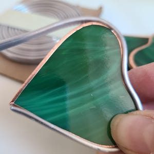 May include: A handcrafted stained glass heart with a malachite green pattern. The glass is framed with a silver-colored metal and copper edging. A roll of metal foil is in the background.