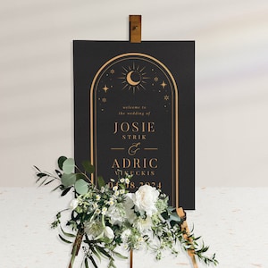 Op de afbeelding: Zwart huwelijkswelkombord met gouden belettering en een maan- en sterrenontwerp. Het bord luidt "Welkom op de bruiloft van Josie Strik & Adric Vinckis 04.08.2024".