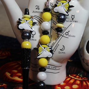 May include: A black pen with a jewelled barrel and a beaded charm featuring yellow, white, and black beads, and a cartoon character. A matching keychain and charm are also visible. The items are displayed against a white hand-shaped ceramic.