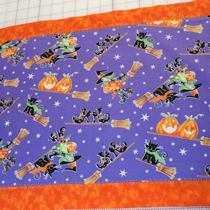 Könnte beinhalten: Halloween-Tischläufer mit einem lila Mittelteil, der Hexen auf Besen, Kürbisse und schwarze Katzen zeigt. Der Rand ist orange. Der Stoff ist mit weißen Sternen gemustert.