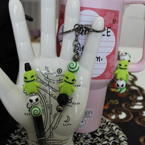May include: A black pen with a green and white swirl charm, a black bead, and a white and black Jack Skellington charm.  The pen is on a white hand with a green and white swirl charm, a black bead, and a green Jack Skellington charm.  The hand is holding a pink tumbler with the text "Can't-E" on it.