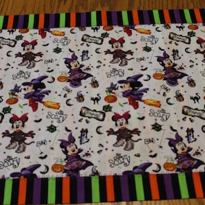 May include: A rectangular fabric table runner featuring a white background with a repeating pattern of Minnie Mouse dressed as a witch, pumpkins, and the text "Be Scary". The runner has a striped border in alternating green, orange, and purple.