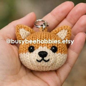 Op de afbeelding: Een gebreide sleutelhanger in de vorm van een Shiba Inu hondenkop van beige en crèmekleurig garen met zwarte plastic ogen en een geborduurde zwarte neus en mond. De tekst @busybeehobbies.etsy staat op het gezicht en een zilveren metalen ring is aan de bovenkant bevestigd.