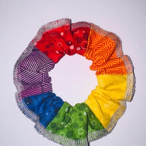 May include: A circular fabric hair scrunchie featuring a rainbow color palette with distinct segments in red, orange, yellow, green, blue, and purple. Each section displays a unique pattern, and the outer edge is finished with a silver-toned serged seam.