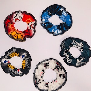 May include: Five fabric hair scrunchies with various patterns and colors. One is red, yellow, and white with a comic book style print. Another is blue and black. A third is dark green and white. A fourth is black, gray, and yellow. The last is white and black.