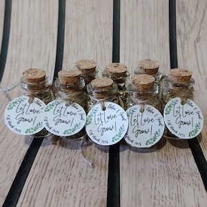Pode incluir: Uma coleção de pequenas garrafas de vidro cheias de uma substância verde, cada uma selada com uma rolha e amarrada com barbante. Cada garrafa tem uma etiqueta redonda que diz "Let Love Grow!" com uma data e um design floral. Dispostas em uma superfície de madeira.