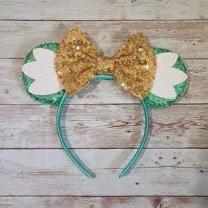 May include: Green and white mouse ears headband with a gold glitter bow. The headband is made of satin and the ears are covered in sequins.