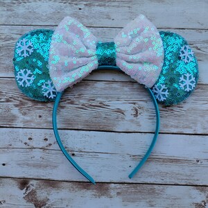 May include: A pair of blue and white sparkly Minnie Mouse ears with a white bow and snowflake embellishments. The headband is blue.
