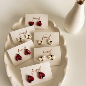 May include: A collection of stud earrings displayed on white cards within a decorative white dish. The earrings feature ladybug and bee designs in red, black, white, and gold. The cards have the word "found" printed on them.