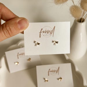May include: A pair of gold-toned dragonfly stud earrings attached to a white display card featuring the text found jewelrySD. The earrings are small with detailed wings and a slender body, presented on a clean white rectangular card.