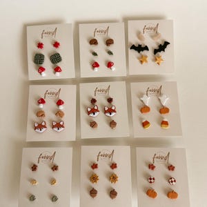 May include: Nine white display cards featuring various pairs of small stud earrings with autumn and Halloween themes, including shapes like foxes, mushrooms, pumpkins, bats, stars, acorns, leaves, and ghosts, each labeled with the word found.