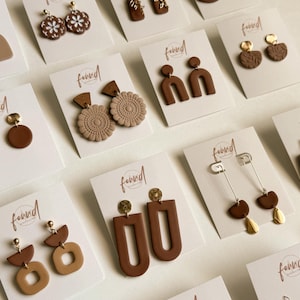 May include: A collection of various polymer clay earrings in shades of brown, tan, and terracotta, displayed on white branded cards. The earrings feature geometric shapes, floral patterns, and gold-toned metal accents, including studs and dangles.