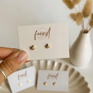 May include: Gold-colored seashell stud earrings displayed on a white card with the brand name "found jewelryNJ". The earrings are presented on a beige plate, with a vase of dried flowers in the background.