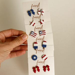 May include: A collection of patriotic stud earrings in red, white, and blue. Designs include bows, stars, flags, and popsicles. The earrings are displayed on white cards with the brand name "found jewelry nY".