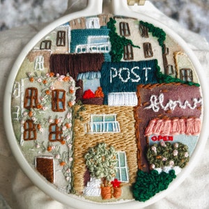 May include: Embroidered hoop art featuring a colorful cityscape with buildings, windows, and greenery. The buildings have signs that read "POST" and "flower" with an "OPEN" sign on a shop.