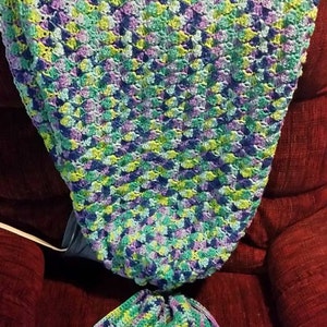 May include: A crocheted mermaid tail blanket in shades of green, blue, purple, and yellow. The blanket is folded in half and has a matching crocheted hat.