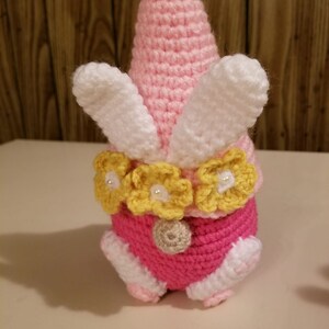 May include: A pink crocheted gnome with white bunny ears and a yellow flower crown. The gnome has a pink and white crocheted body and a brown crocheted nose.