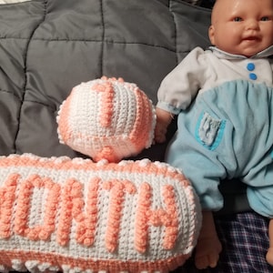 May include: A crocheted pillow with the word "MONTH" in pink and white yarn. A smaller crocheted ball with the number "1" is in the background. A baby doll is sitting on a plaid blanket.