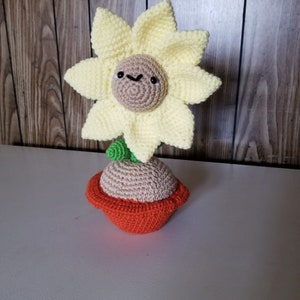 May include: A crocheted sunflower with a brown face and a happy expression, standing in a small orange pot. The sunflower is yellow and the pot is orange.