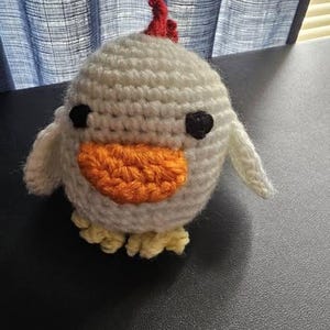 May include: A handmade crochet chick toy. The chick is primarily white with an orange beak, black eyes, a red comb, and yellow feet. The wings are small and white. The toy is sitting on a black surface.