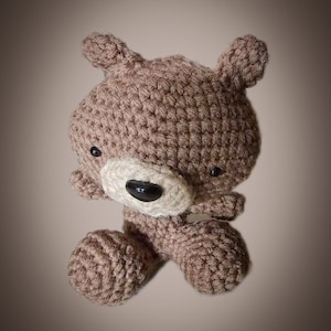 Op de afbeelding: Een bruine gehaakte teddybeer met zwarte ogen en een witte neus. De beer zit met gekruiste benen.