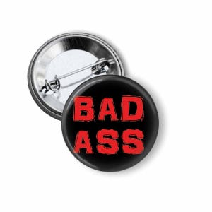 May include: A black button with the words "BAD ASS" in red, bold letters. The button is round and has a metal backing with a pin for attachment. The button is a novelty item.