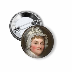 May include: A round button pin featuring a portrait of a woman in a white bonnet with lace trim. The portrait has a brown background and the woman has rosy cheeks. The pin has a silver metal backing.