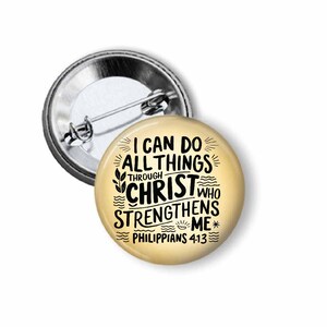 May include: A round button pin with a metallic silver backing and a cream-colored face. The face features black text that reads "I CAN DO ALL THINGS THROUGH CHRIST WHO STRENGTHENS ME" and "PHILIPPIANS 4:13".
